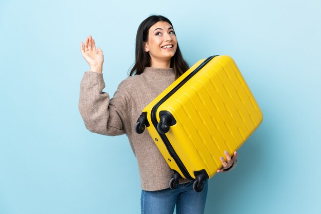 Jong donkerbruin meisje over geïsoleerd blauw in vakantie met reiskoffer en het groeten