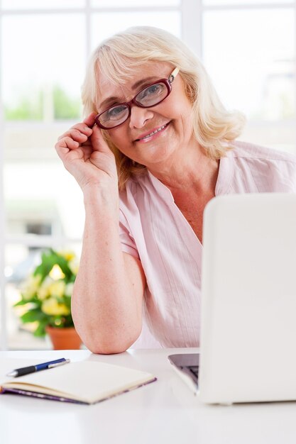 Jong en actief voelen. Vrolijke senior vrouw die haar bril aanpast en glimlacht terwijl ze op laptop werkt