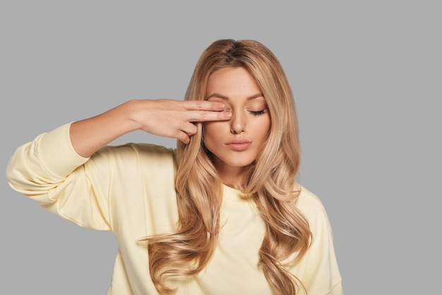 Jong en mooi. Aantrekkelijke jonge vrouw die oog bedekt met vingers terwijl ze tegen een grijze achtergrond staat