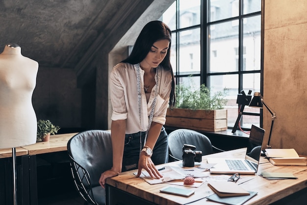 Jong en slim. mooie jonge vrouw met meetlint om haar nek die naar modetijdschrift kijkt terwijl ze op het bureau in haar atelier leunt