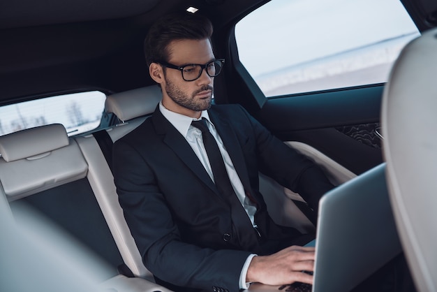 Jong en succesvol. Knappe jongeman in volledig pak aan het werk met laptop terwijl hij in de auto zit