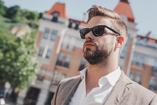 Jong en succesvol portret van een bruinharige man die een zonnebril draagt en wegkijkt in de stad