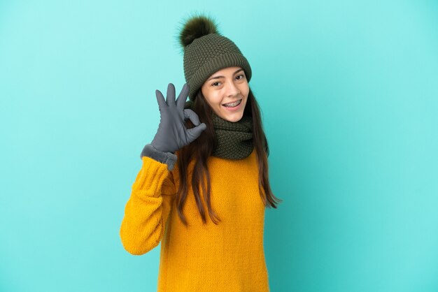 Jong Frans meisje geïsoleerd op blauwe achtergrond met winter hoed weergegeven: ok teken met vingers