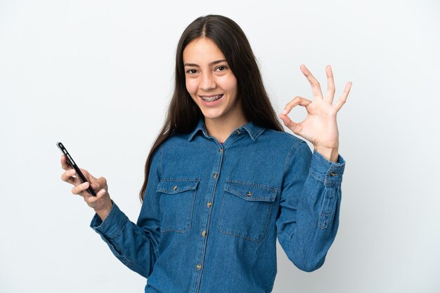 Jong Frans meisje geïsoleerd op een witte achtergrond met behulp van mobiele telefoon en doen OK sign