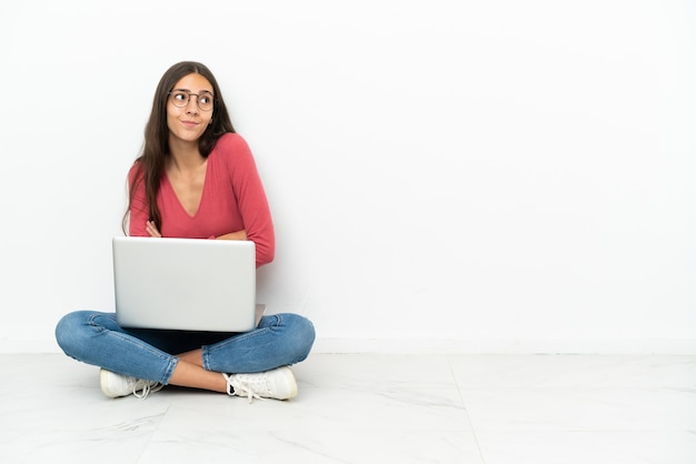 Jong Frans meisje zittend op de vloer met haar laptop twijfels gebaar maken terwijl het opheffen van de schouders