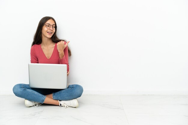 Jong frans meisje zittend op de vloer met haar laptop wijzend naar de zijkant om een product te presenteren