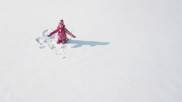 Jong geitje dat op een sneeuw een sneeuwengel maakt. Luchtfoto van bovenaf. Ze draagt rode kleren