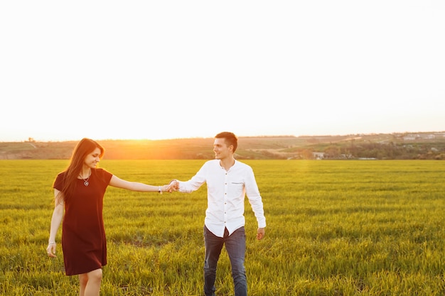 Jong, gelukkig, liefdevol paar, bij zonsondergang, staande in een groen veld, tegen de hemel hand in hand, en genieten van elkaar