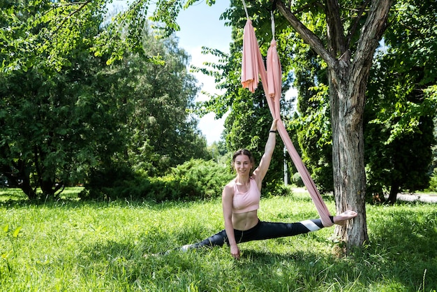 Jong gelukkig meisje traint met vliegyoga buiten in de zomer in een linnen hangmat onder een boom Buiten zomeryoga vliegconcept