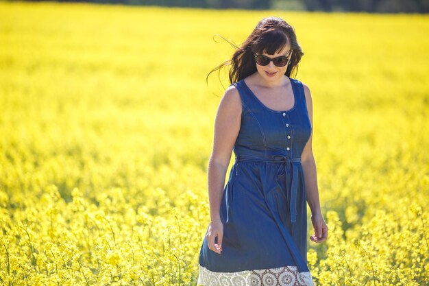 Jong gelukkig mooi groot model meisje in blauwe jurk en zonnebril op bloeiend koolzaadveld in de zomer