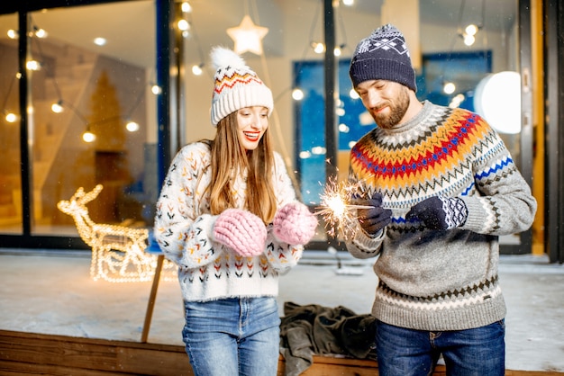 Jong gelukkig stel gekleed in truien die wintervakantie vieren met Bengaals vuur voor een prachtig versierd huis