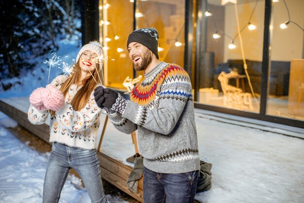 Jong gelukkig stel gekleed in truien die wintervakantie vieren met bengaals vuur voor een prachtig versierd huis