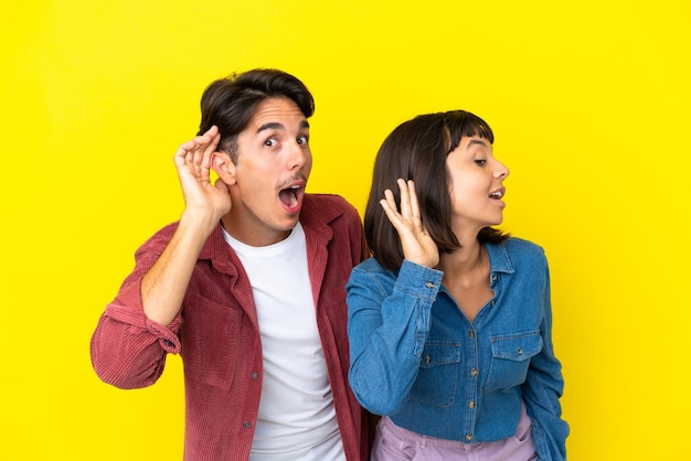 Jong gemengd ras paar geïsoleerd op gele achtergrond luisteren naar iets door hand op het oor te leggen