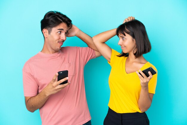 Jong gemengd ras paar met mobiele telefoon geïsoleerd op blauwe achtergrond twijfels tijdens het hoofd krabben