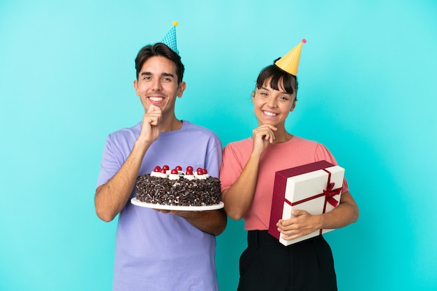 Jong gemengd ras paar met verjaardagstaart en cadeau geïsoleerd op blauwe achtergrond glimlachend met een zoete uitdrukking