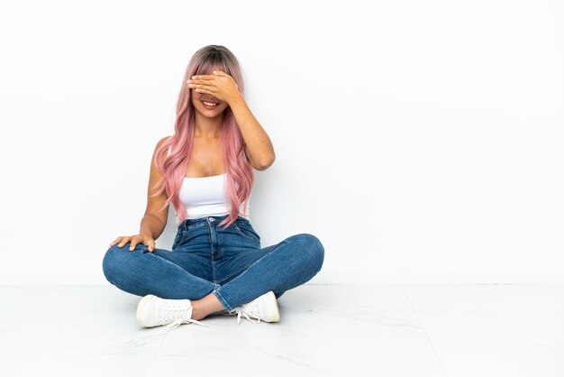 Jong gemengd ras vrouw met roze haren zittend op de vloer geïsoleerd op een witte achtergrond die ogen bedekt door handen. Wil je iets niet zien