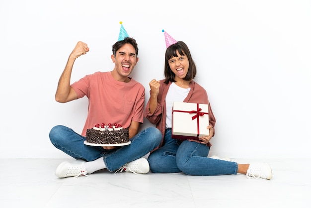 Jong gemengd raspaar dat een verjaardag viert zittend op de vloer geïsoleerd op een witte achtergrond die een overwinning viert