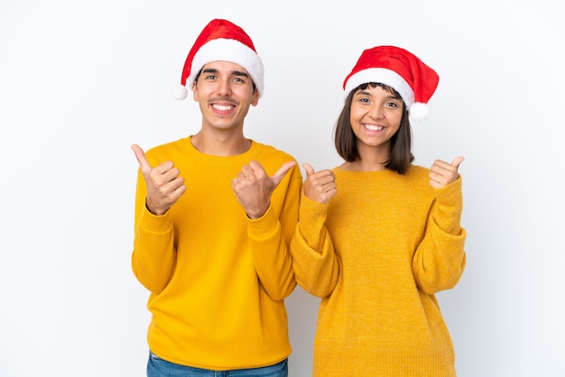 Jong gemengd raspaar dat Kerstmis viert dat op witte achtergrond wordt geïsoleerd en een duim omhoog gebaar geeft met beide handen en glimlacht