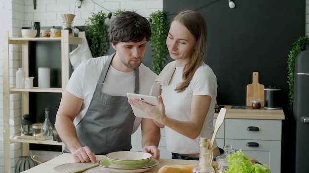 Jong getrouwd stel maakt gebruik van een digitale tablet en glimlacht tijdens het koken in de keuken thuis