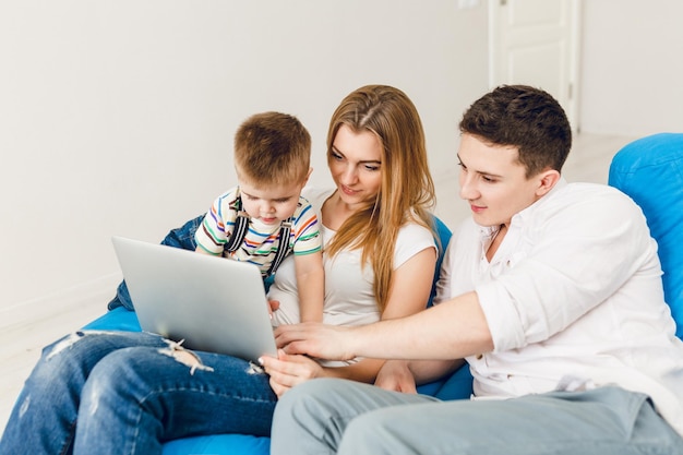 Jong gezin van twee ouders en een jongenskind zitten op blauwe zakstoelen. Ze dragen een wit t-shirt en een spijkerbroek. Moeder houdt een laptop vast en een jongen speelt erop. Ze zitten in een studio met witte muren.