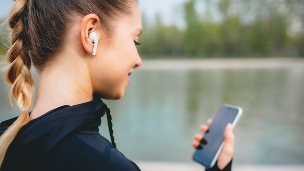 Jong glimlachend meisje dat sport maakt en in het park rent met haar telefoon om de muziek te luisteren met een draadloze koptelefoon op zonsondergang in de stad die naar het scherm kijkt