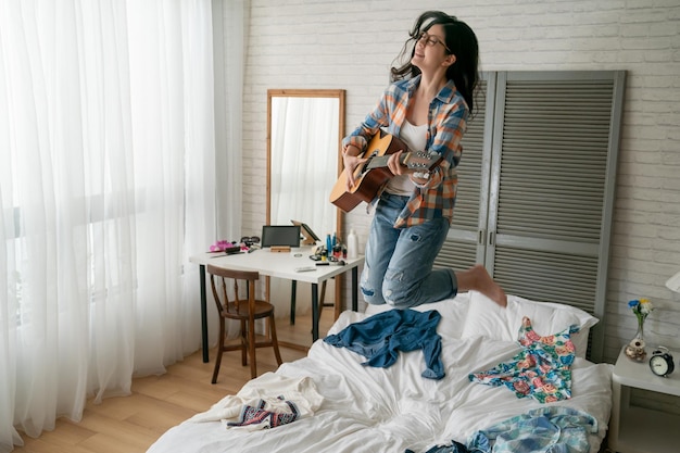 jong grappig meisje akoestische gitaar spelen veel plezier dansen op bed thuis. vrolijke dame die in de slaapkamer springt geniet van muziek die liedjes zingt in het appartement met rommelige kleurrijke kleding. gelukkige vrouw die lacht