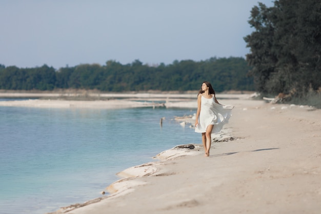 Jong heel mooi meisje met lang haar in een witte jurk aan het meer. Het meisje geniet van de rest.