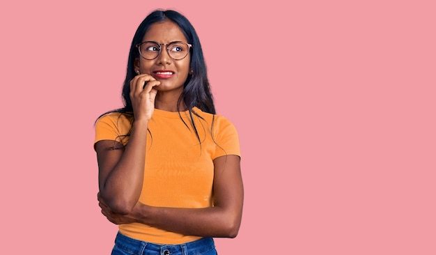 Jong indisch meisje dat casual kleding en een bril draagt, ziet er gestrest en nerveus uit met handen op de mond bijtende nagels angstprobleem