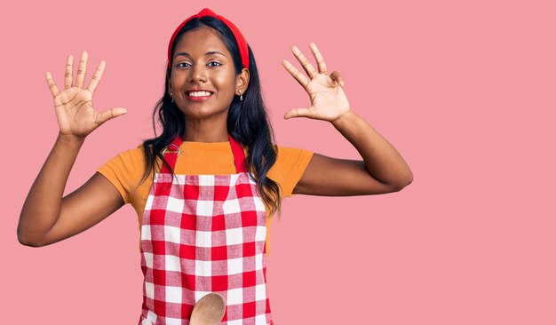 Jong indisch meisje dat een professionele bakkersschort draagt en met vingers nummer negen naar boven wijst terwijl ze zelfverzekerd en gelukkig glimlacht.