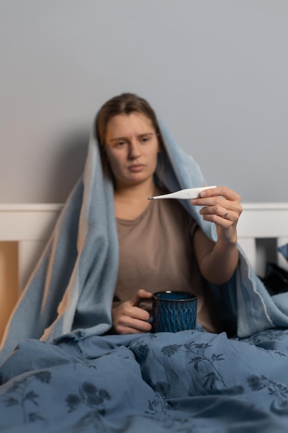 Jong Kaukasisch meisje bedekt met een deken zittend in bed en in de hand witte thermometer te houden. Vrouw heeft symptomen van verkoudheid en griep. Vrouw die lijdt aan hitte en zwakte. Gezondheidszorgconcept.