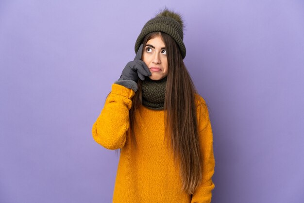 Jong Kaukasisch meisje dat met de winterhoed op purpere achtergrond wordt geïsoleerd die een idee denkt