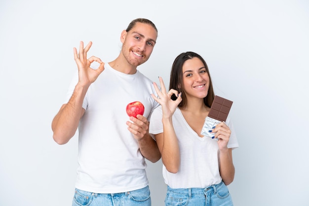 Jong Kaukasisch paar met chocolade en appel geïsoleerd op een witte achtergrond met een ok teken met vingers