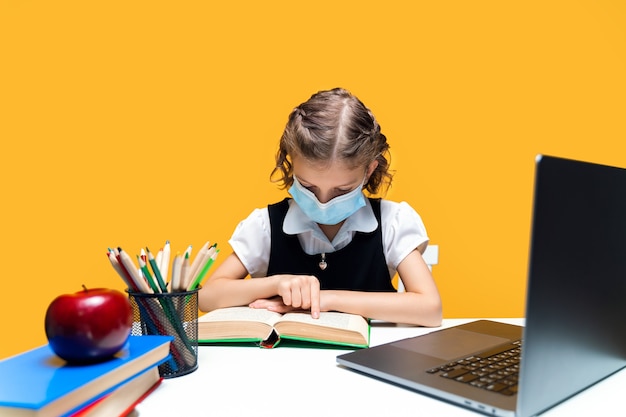 Jong Kaukasisch schoolmeisje zit aan de balie leesboek in masker met laptop afstandsonderwijs