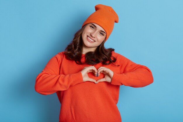 Jong Kaukasisch wijfje dat oranje sweater en hoed draagt die hartgebaar met handen maakt en direct camera met gelukkige uitdrukking bekijkt, die over blauwe muur wordt geïsoleerd.