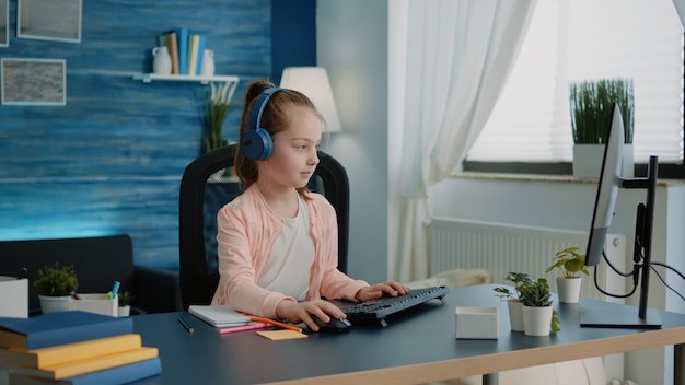 Jong kind tekenen op leerboek en computer gebruiken terwijl het dragen van een koptelefoon voor online les op afstand. Schoolmeisje dat kleurrijke potloden gebruikt om op een notitieboekje te tekenen en op het toetsenbord te typen.