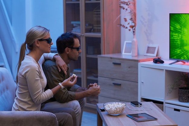 Jong koppel 3D-bril zittend op de bank popcorn eten en kijken naar film in een donkere kamer thuis