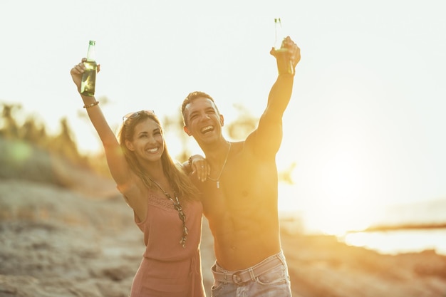 Jong koppel dansen en bier drinken op het strand aan zee. Genieten in prachtige zonsondergang en hun liefde.