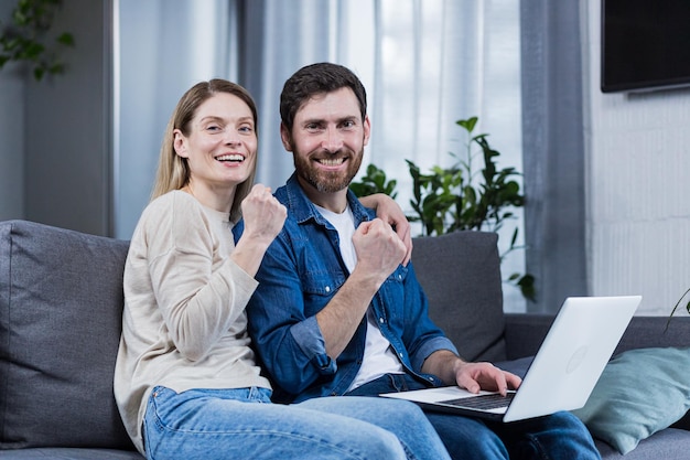 Jong koppel familie man en vrouw zittend op de bank thuis met behulp van laptop iets winnen en goed nieuws krijgen blij lachend knuffelen