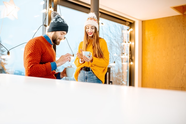 Foto jong koppel gekleed in heldere truien en hoeden staan samen met warme dranken bij de open haard in het moderne huis tijdens de winter