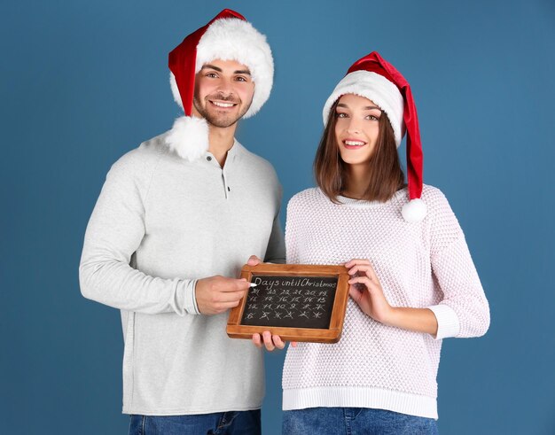 Jong koppel in kerstmutsen met schoolbord dagen tellen tot Kerstmis, op kleur achtergrond