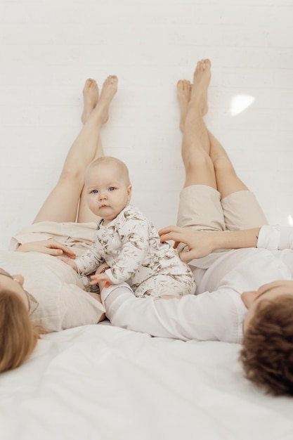 Jong koppel in witte kleren liggend met benen rechtop hoog op bed met baby baby sittin