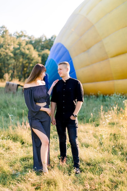 Jong koppel in zwarte kleding, knuffelen en hand in hand, terwijl staande in zomer veld voor kleurrijke luchtballon, de vlucht voorbereiden