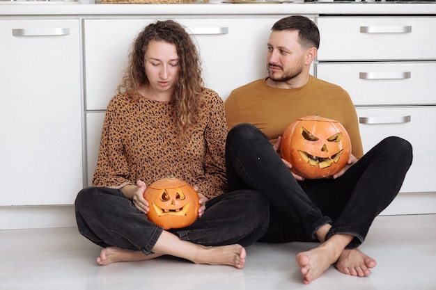 Jong koppel man en vrouw zittend op de vloer in de keuken thuis met plezier en voorbereiding voor halloween praten vrolijk lachen.