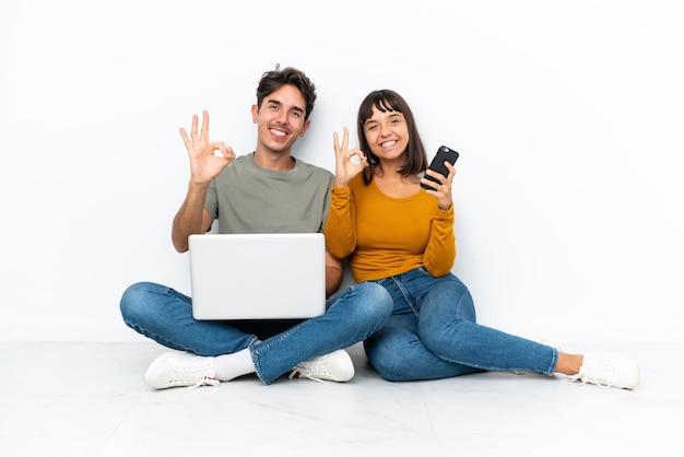 Jong koppel met een laptop en mobiel zittend op de vloer met een ok teken met vingers