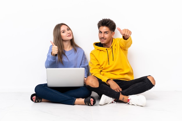 Jong koppel met een laptop zittend op de vloer op wit met duimen omhoog omdat er iets goeds is gebeurd