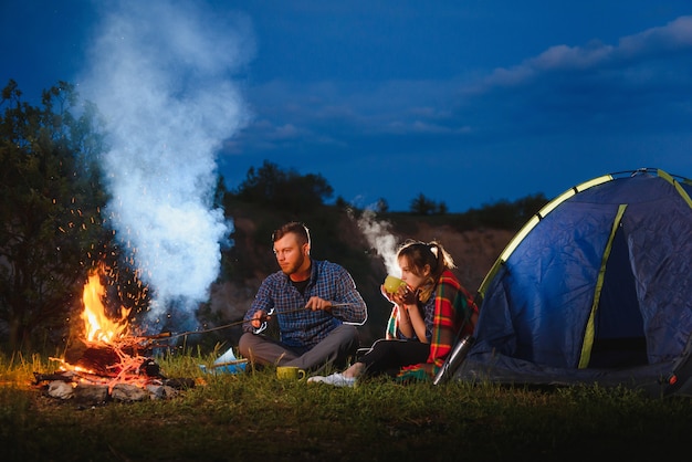 Jong koppel met een rust bij vuur naast kamp en blauwe toeristische tent, het drinken van thee, genieten van de nachtelijke hemel.