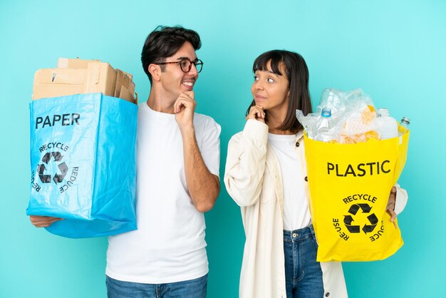 Jong koppel met een zak vol plastic en papier om te recyclen geïsoleerd op een blauwe achtergrond terwijl ze naar elkaar kijken