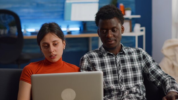 Jong koppel tussen verschillende rassen chatten op video-oproepconferentie in de woonkamer. Multi-etnische mensen praten op laptopcamera voor communicatie op afstand terwijl ze thuis op de bank zitten.