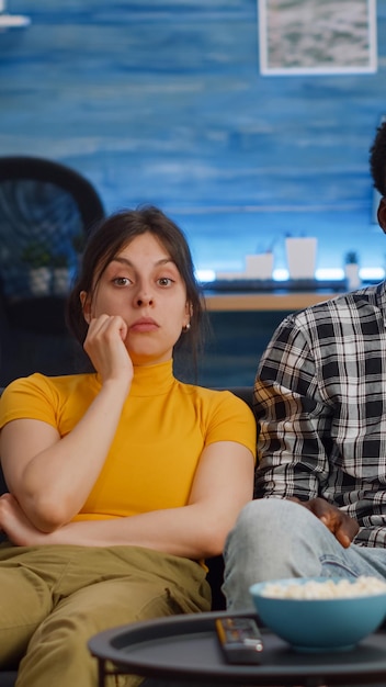 Jong koppel tussen verschillende rassen televisie kijken popcorn eten in de woonkamer. Getrouwde multi-etnische mensen die naar de camera kijken en een snack hebben die genieten van ontspannende activiteiten. Liefhebbers van gemengd ras