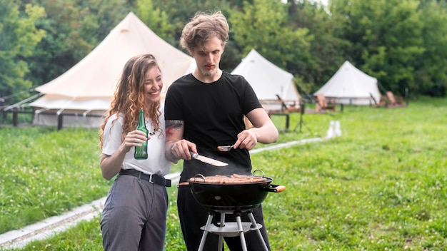 Jong koppel vlees bakken op de grill en bier drinken tenten op de achtergrond